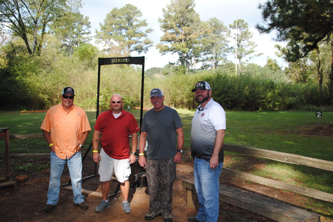 Four men standing outdoors near a wooden structure with greenery in the background.