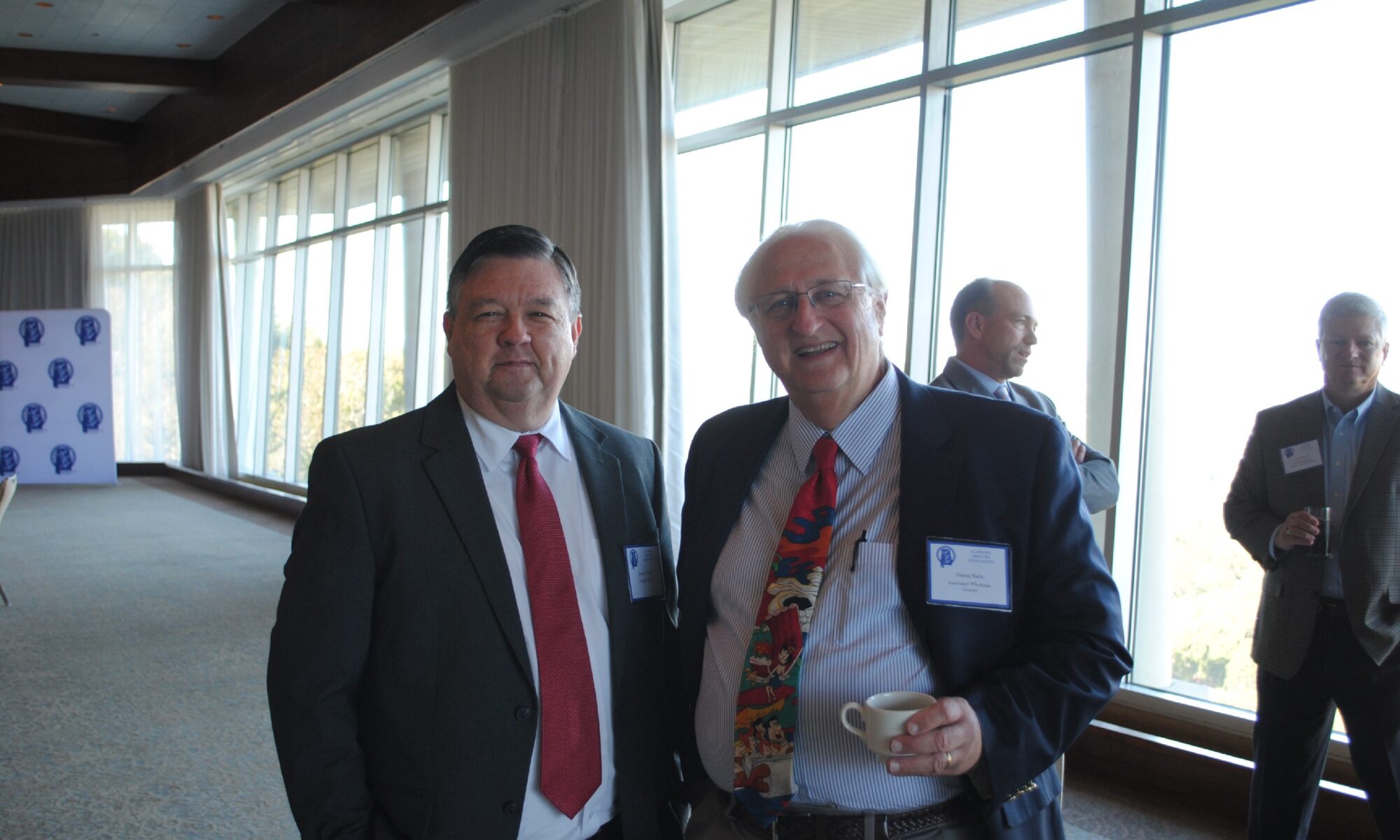 Two men in suits standing indoors near large windows.