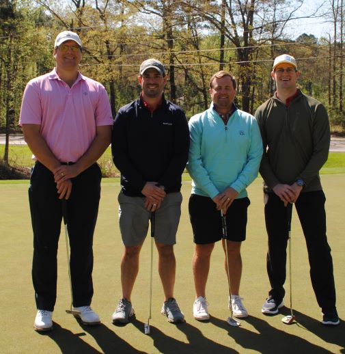 Four men standing together on a golf course on a sunny day.